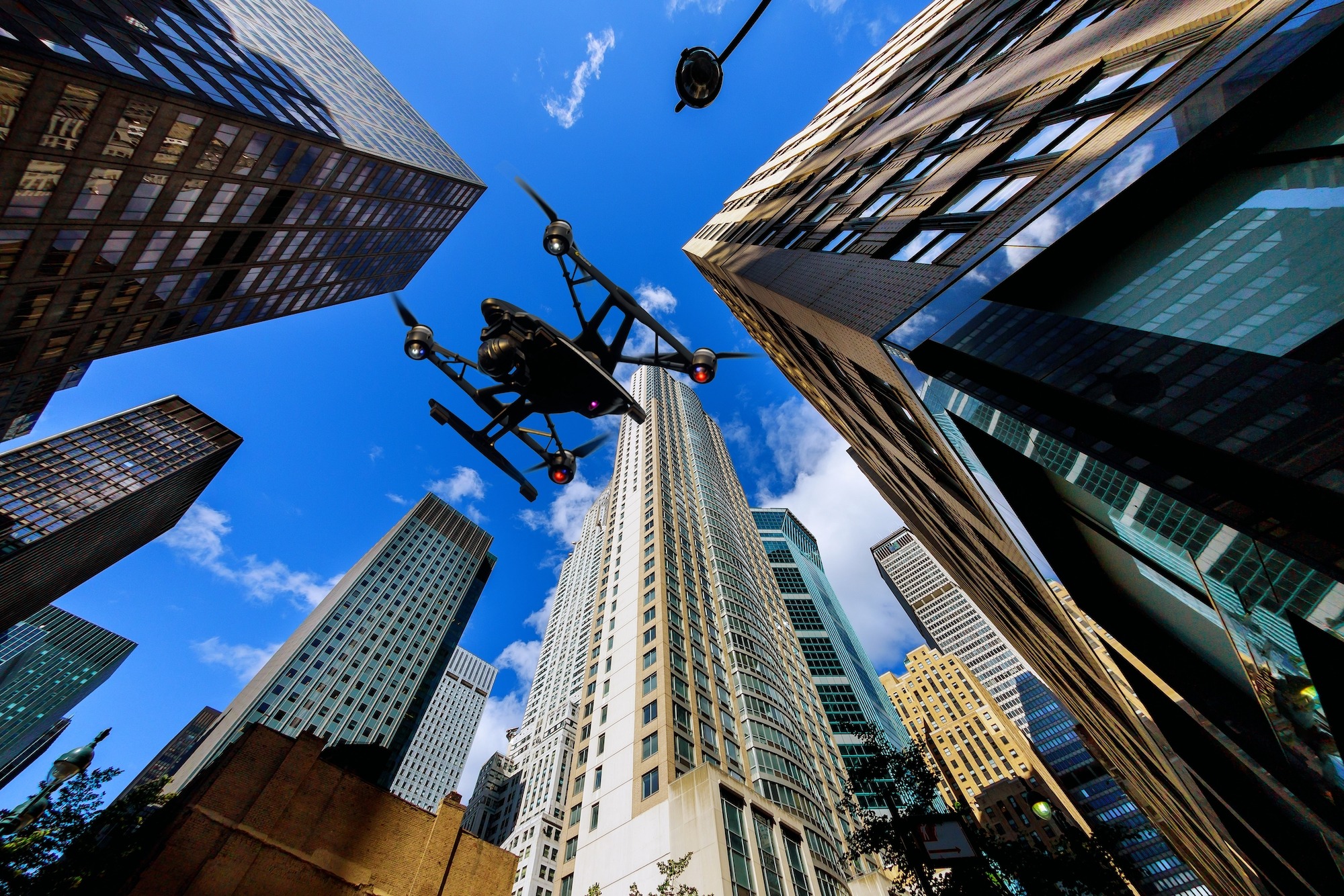 A drone hovers above a cityscape, surrounded by tall skyscrapers under a blue sky, highlighting urban innovation and technology.