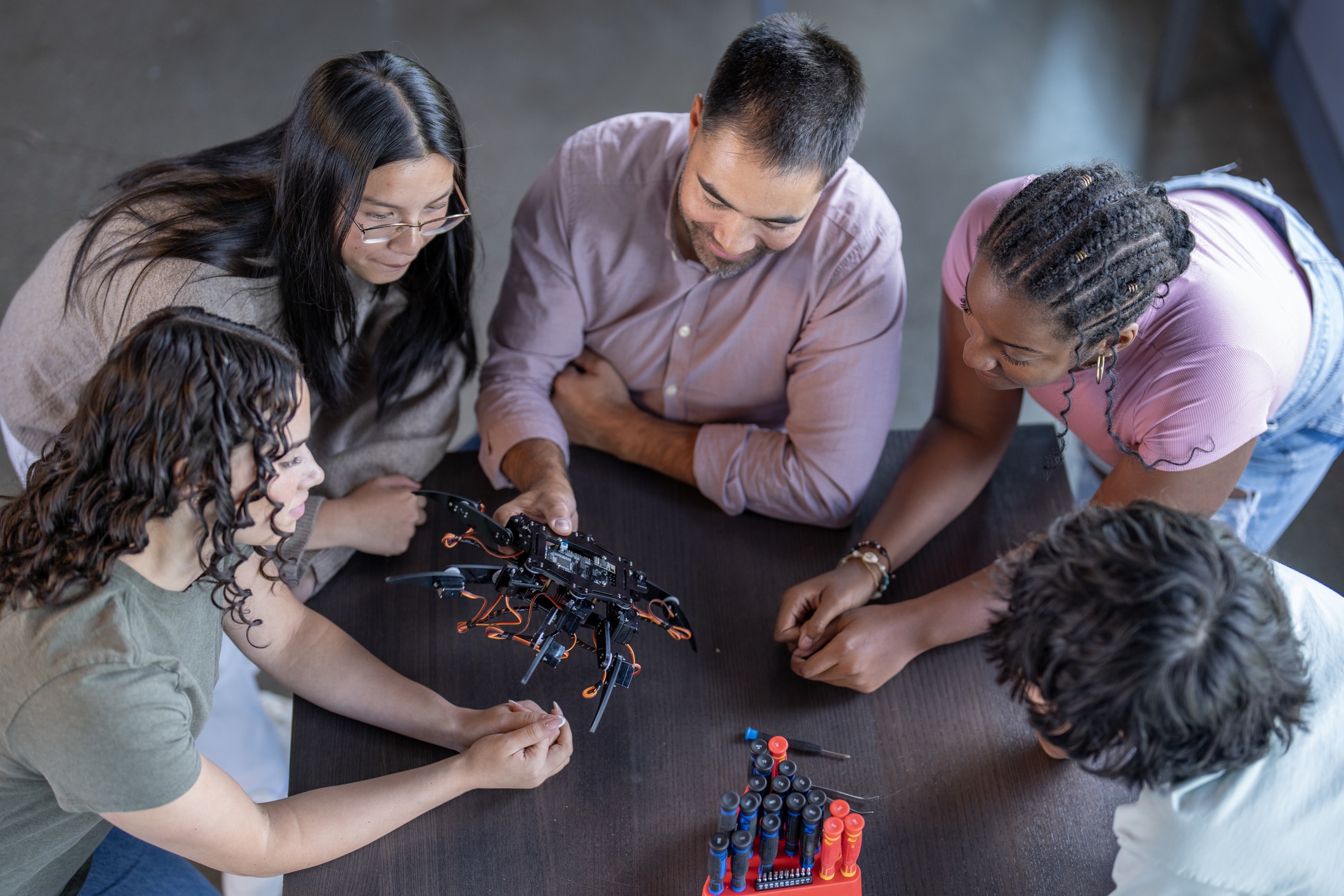 A small group of secondary students huddle around their teacher as he holds out a robot and they explore the project together. The students are each dressed casually and appear very enthralled with the robot.