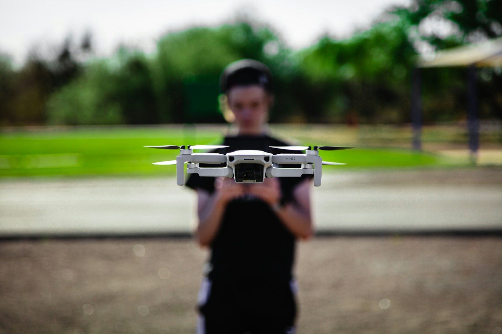 A white Mini 2 drone hovers in focus, with a person in the background controlling it. This highlights drone operation in an outdoor setting.